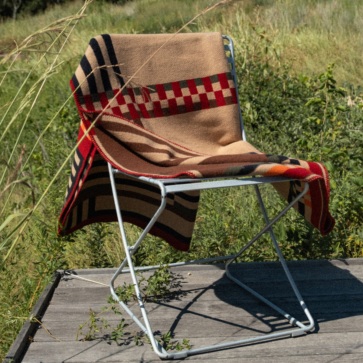 A silver metal folding chair draped with a tan woven blanket featuring red, black, and cream geometric patterns, set outdoors on a wooden deck with tall grass and greenery in the background.