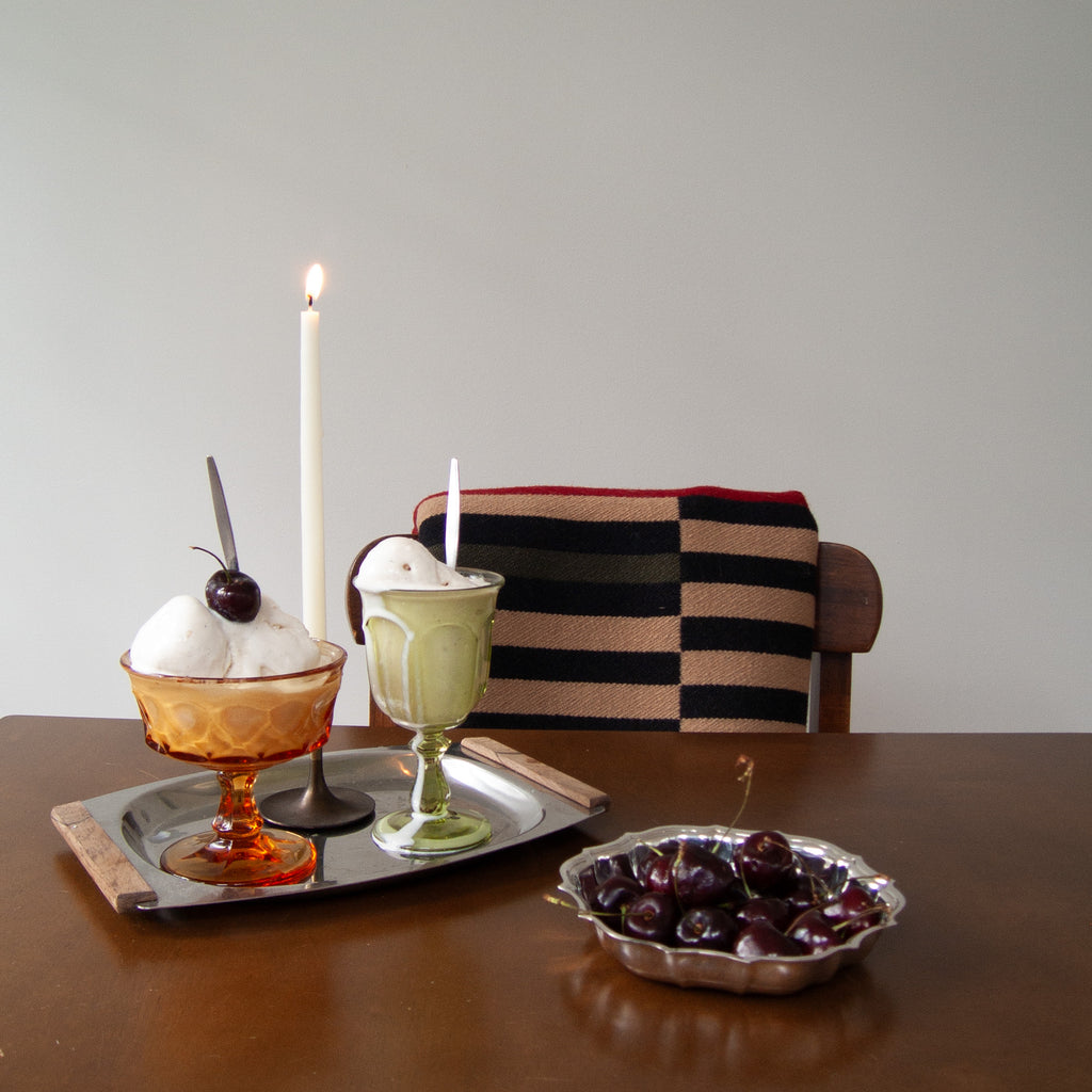 Two dessert glasses on a metal tray on a wooden table, with a lit white candle behind them and a striped cushion on a wooden chair. A bowl of dark cherries sits in the foreground against a plain light gray wall.