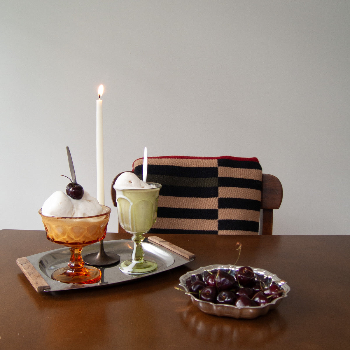 Two dessert glasses on a metal tray on a wooden table, with a lit white candle behind them and a striped cushion on a wooden chair. A bowl of dark cherries sits in the foreground against a plain light gray wall.