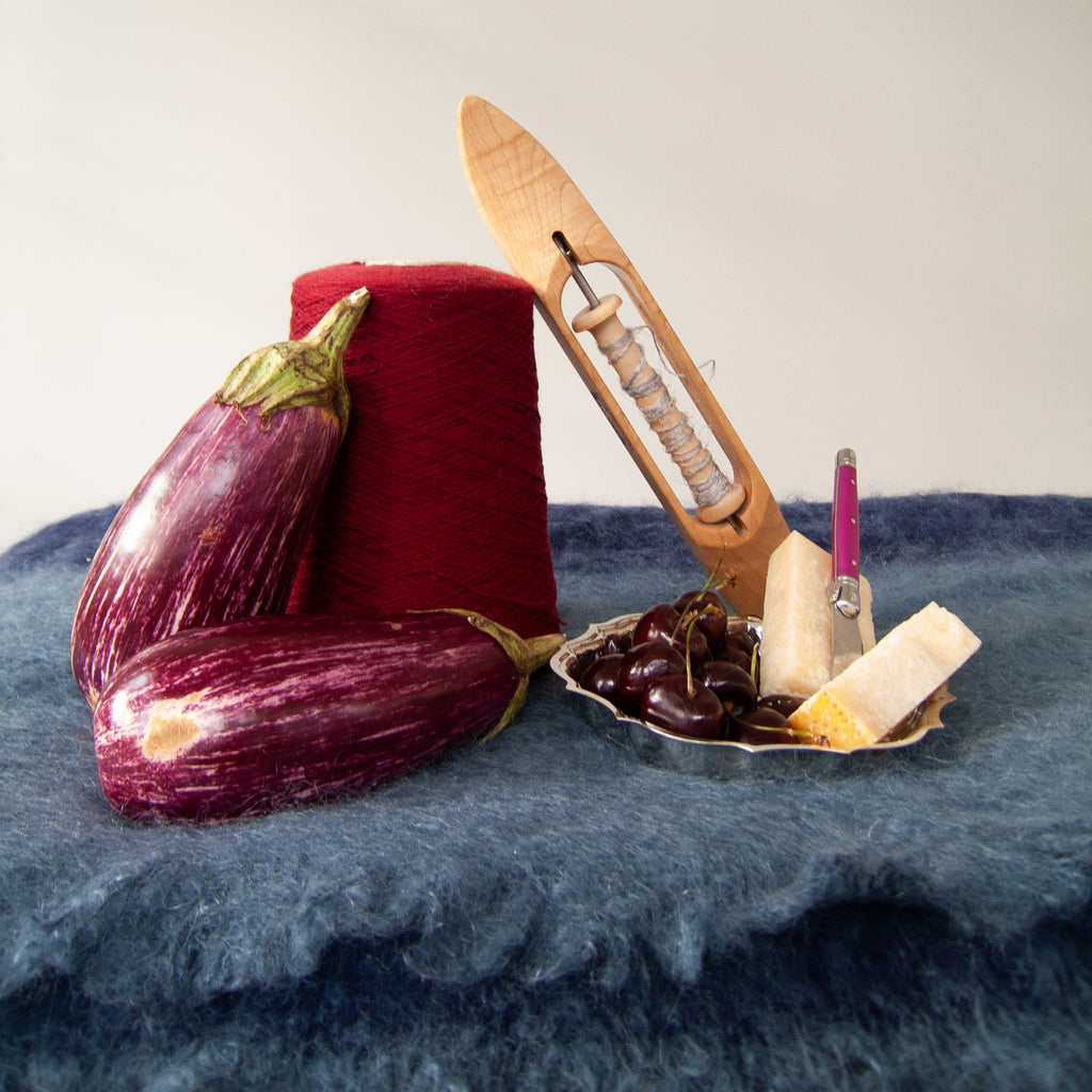 A styled still life featuring a large cone of deep red cashmere yarn, two purple eggplants, a wooden weaving shuttle, a small dish with dark cherries, parmesan, and a small purple cheese knife, arranged on a slate fuzzy mohair blanket.