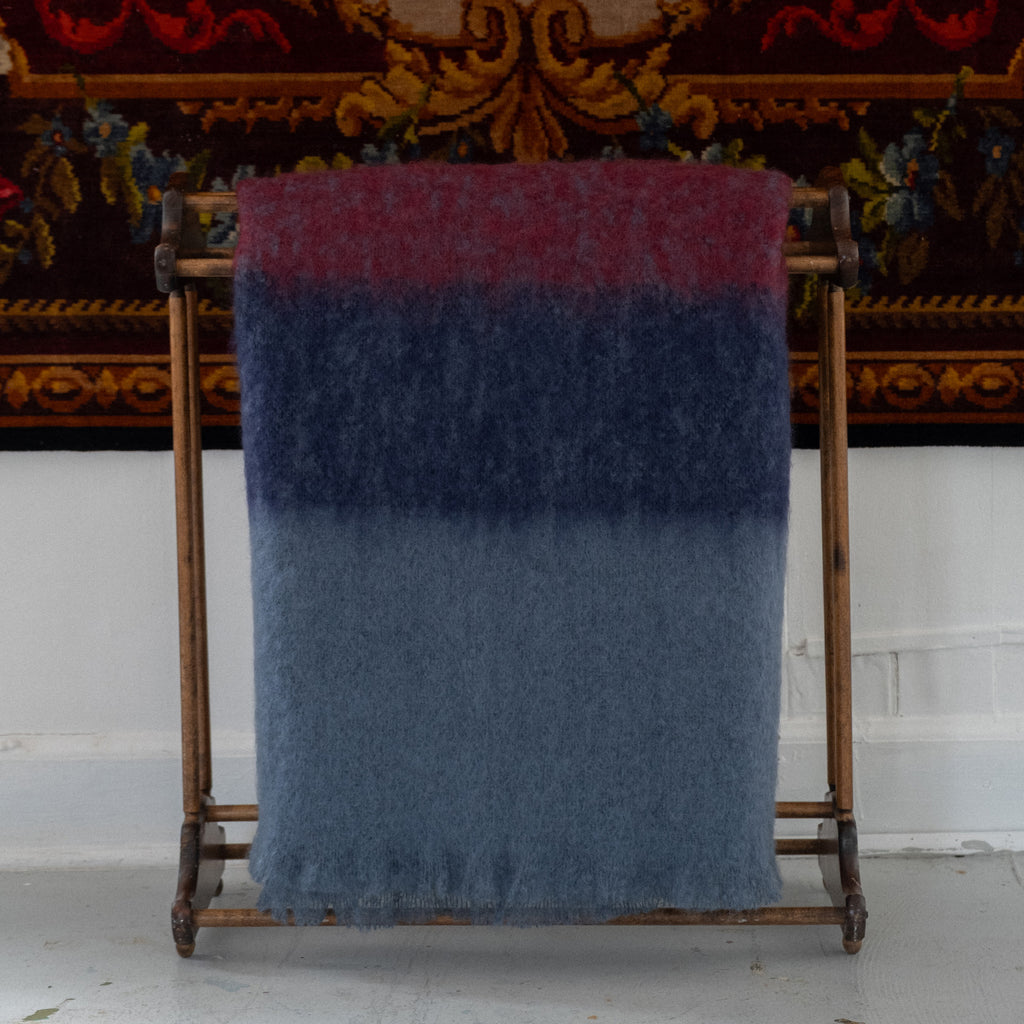 A fuzzy, shaggy throw blanket with a vertical color-blocked pattern in burgundy, dark navy blue, and slate blue, draped over a rustic wood rack with a vintage tapestry in the back