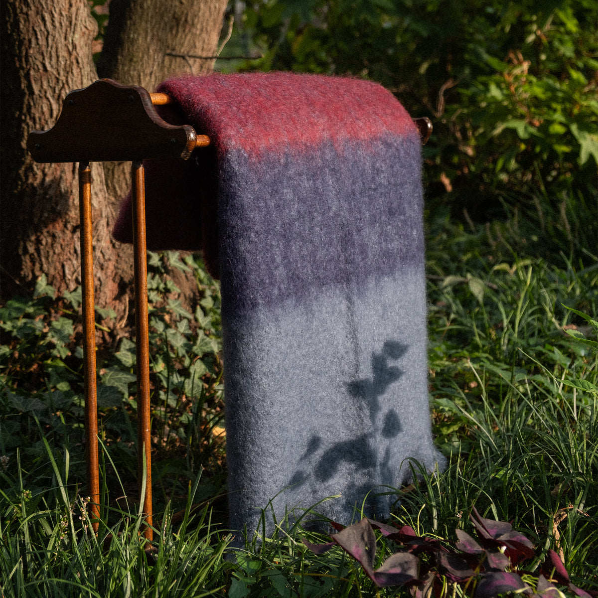 A thick, fuzzy mohair blanket with a vertical color-blocked pattern in navy, burgundy, and slate blue-gray, draped over a rustic wooden stand in an outdoor grassy setting.