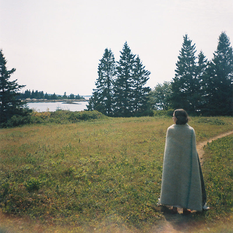 A woman seen from behind standing on a dirt path in a grassy field by the ocean, wearing a long moss colored moahir throw blanket, with evergreen trees  under a pale sky.