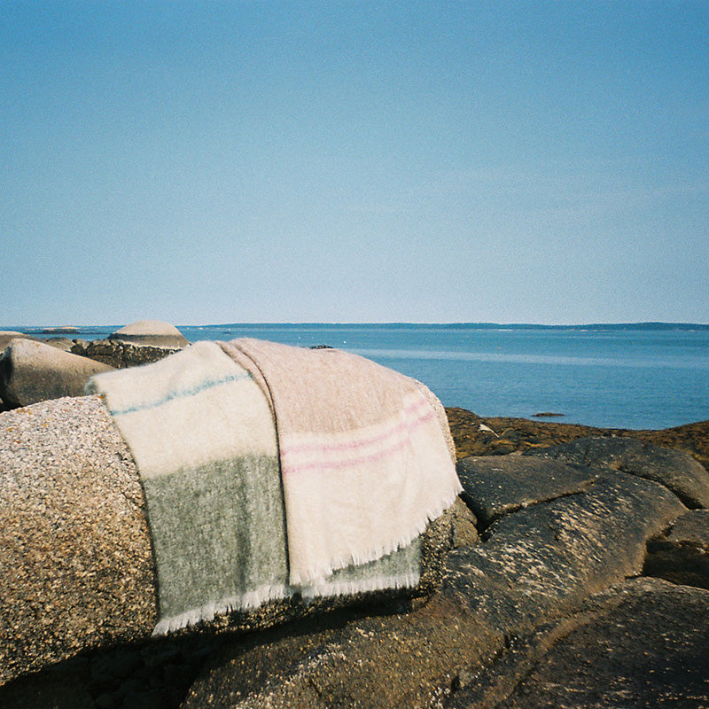 A striped mohair blanket with fringed edges draped over a large rock in an outdoor coastal setting, with ocean water and a clear blue sky in the background.