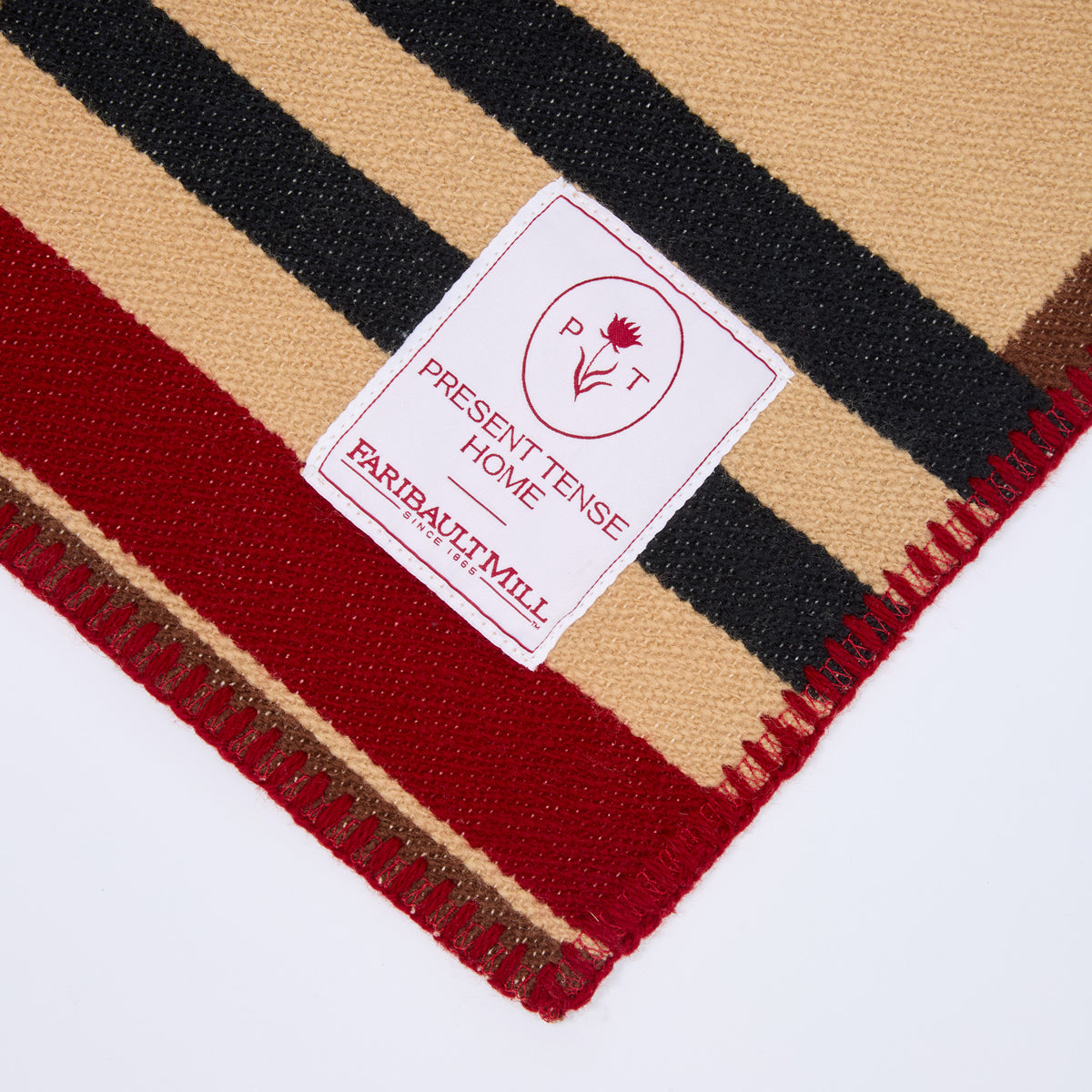 A close-up of a striped woven textile, likely a blanket or throw, with beige, black, and red stripes and a white sewn-in label reading 'PRESENT TENSE HOME' and 'FARBAUT MILL'.