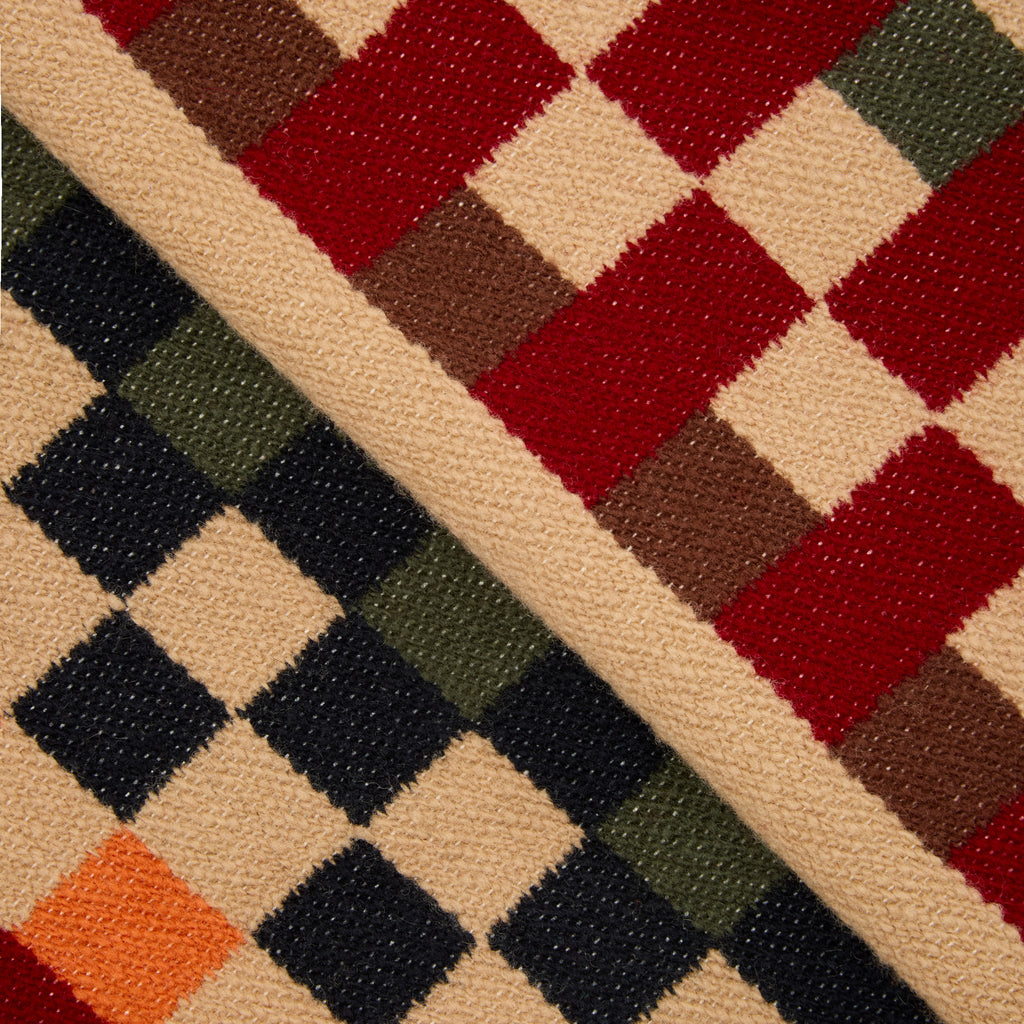Close-up of a woven textile with a geometric check pattern in red, beige, brown, green, black, and orange, divided diagonally into two colorways.