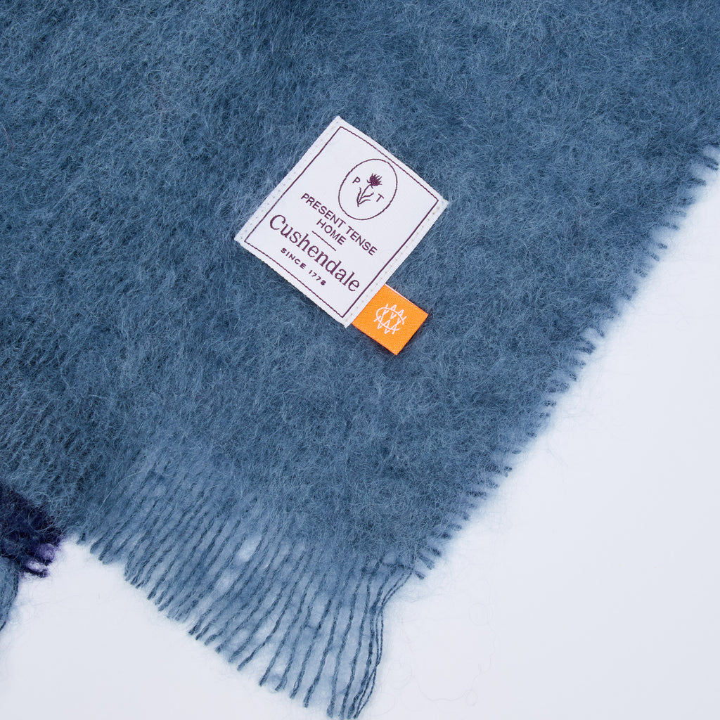 A close-up of a dusty slate blue fuzzy scarf with fringed edges, featuring a white woven brand label that reads 'Present Tense Home Cushendale' and a small orange tag.