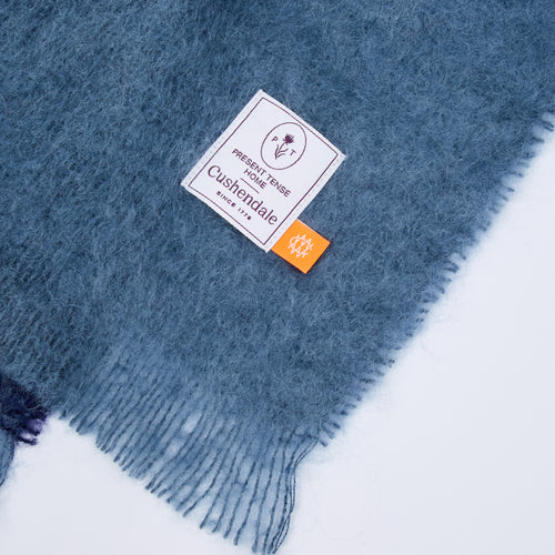 A close-up of a dusty slate blue fuzzy scarf with fringed edges, featuring a white woven brand label that reads 'Present Tense Home Cushendale' and a small orange tag.