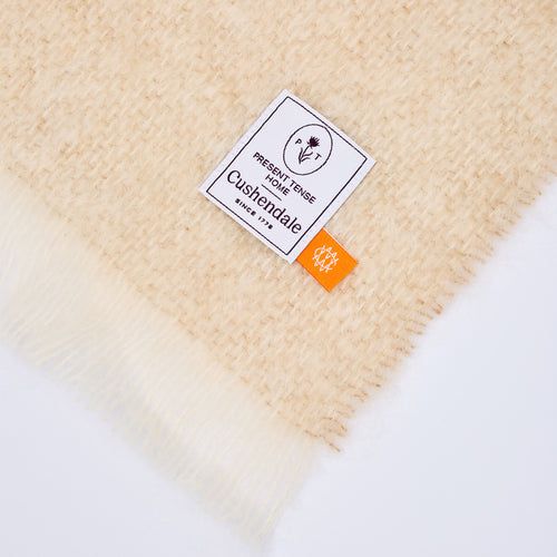 A close-up of a cream-colored, textured woven blanket with fringed edges, featuring a white brand label that reads 'Present Tense Home and Custendale' and an orange tag.