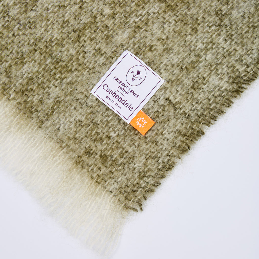 Close-up of a plush, textured throw blanket with a woven pattern in olive green and cream, featuring a white brand label that reads 'Present Tense Home' and a small orange tag.