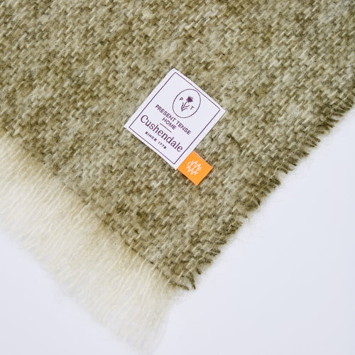 Close-up of a plush, textured throw blanket with a woven pattern in olive green and cream, featuring a white brand label that reads 'Present Tense Home' and a small orange tag.
