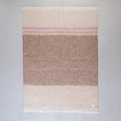 A rectangular mohair throw blanket with horizontal stripes in cream, light brown, and dusty pink, finished with fringed edges, laid flat on a light gray wooden floor.