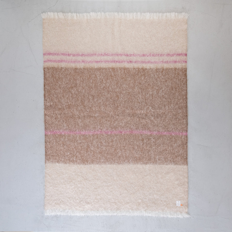 A rectangular mohair throw blanket with horizontal stripes in cream, light brown, and dusty pink, finished with fringed edges, laid flat on a light gray wooden floor.