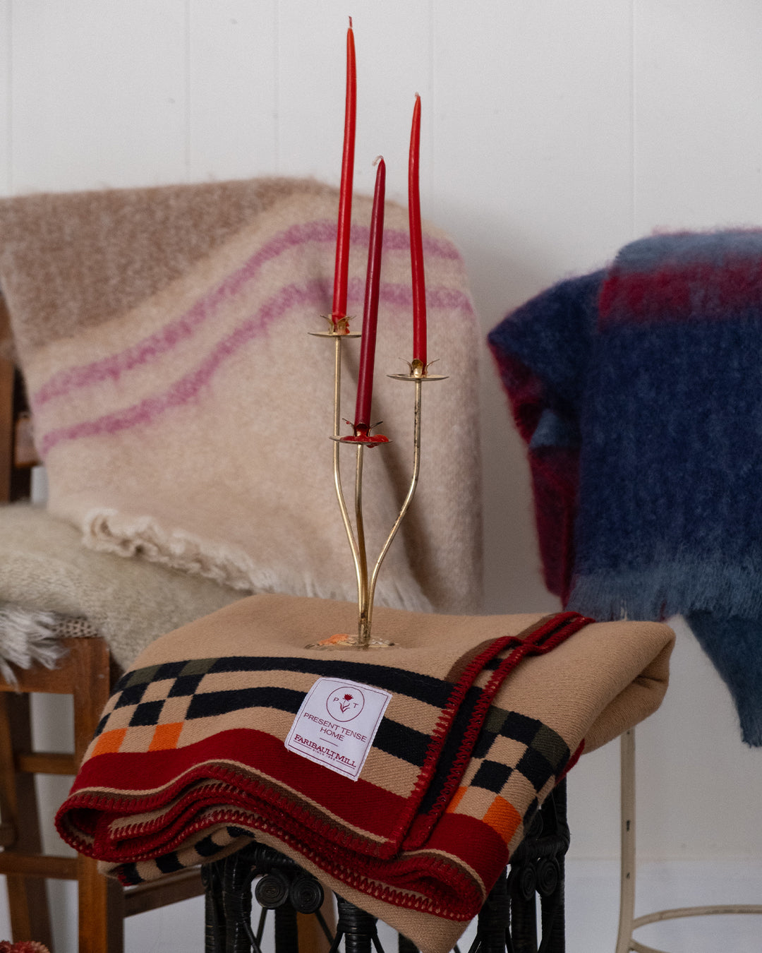 A folded beige wool blanket with a red, black, green, and orange check pattern and a white brand label sits on a table. A brass three-candle holder with red taper candles is placed on top, with two cream wool throws and a blue wool blanket in the background.