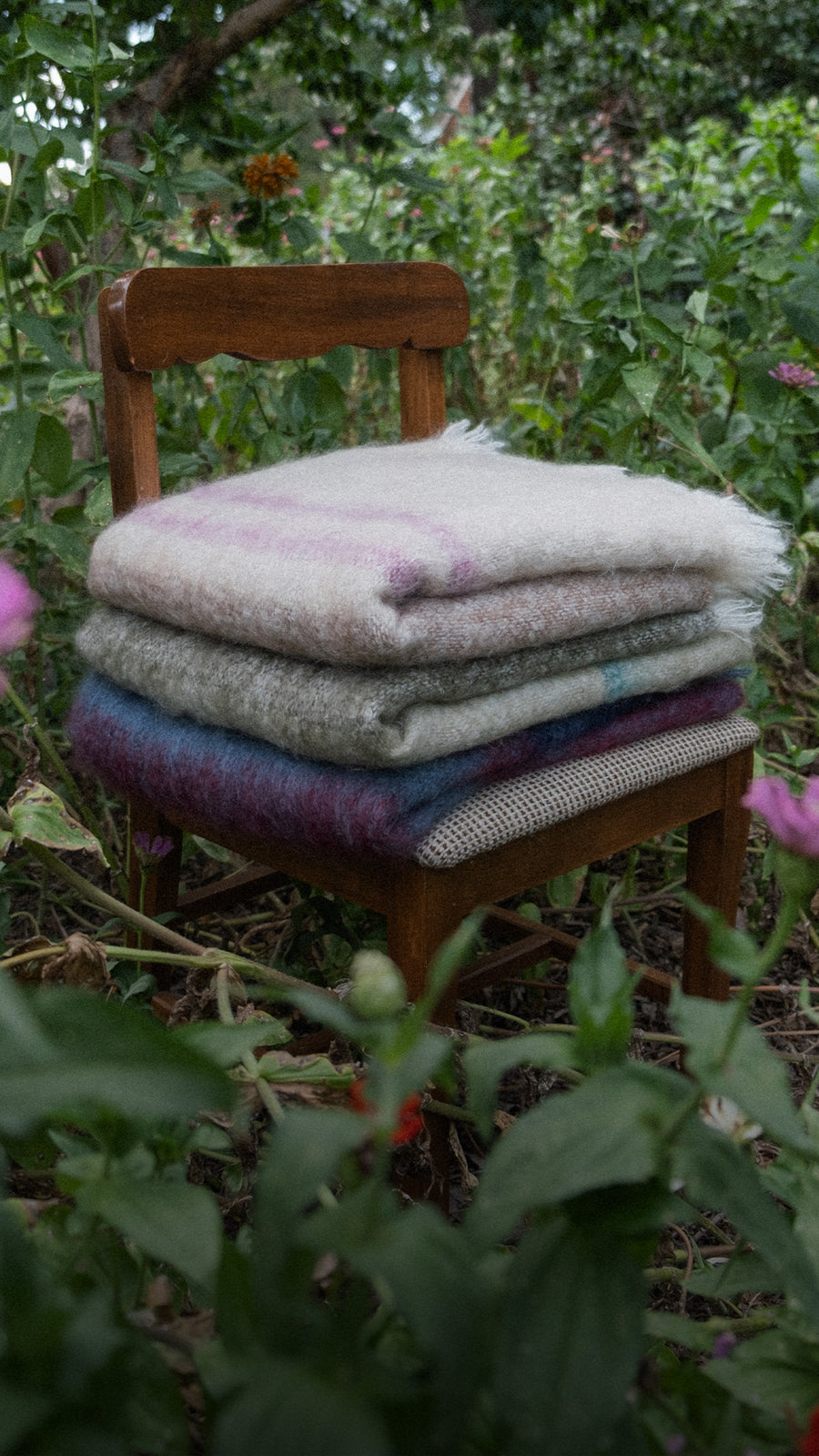A stack of three folded mohair and wool blankets in various colors and patterns sits on a wooden chair placed outdoors in a garden setting with green foliage and flowers.
